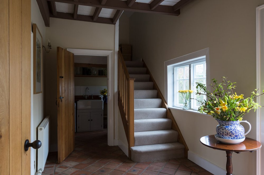  dark unlit  farmhouse hallway with red tile flooring, cream walls, glimpse into under stairs utility, grey carpet stairs 