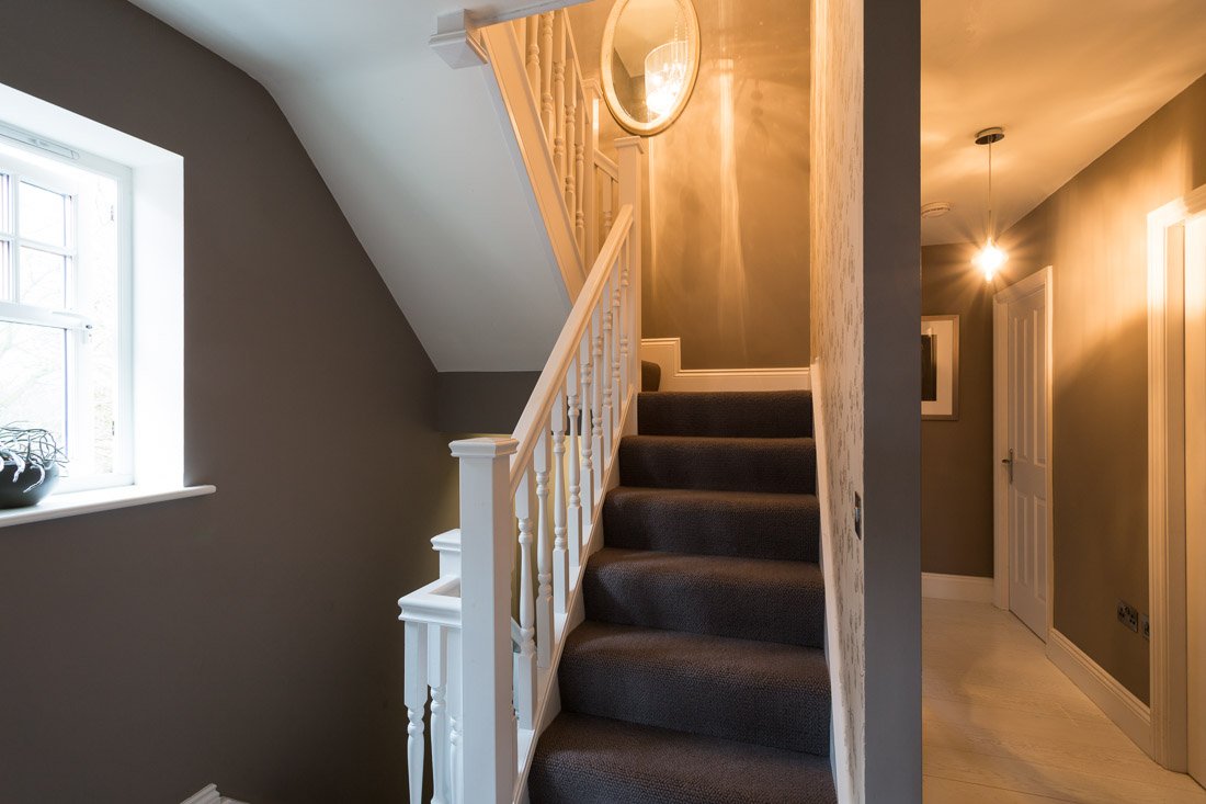  badly lit hallway with stairs going up and down, grey walls, white carpet, grey stair runner, ceiling light casting orange glow 