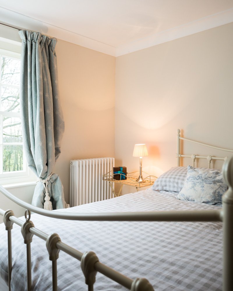  detail looking across bed towards bedside table with tall sash window and blue draped curtains  