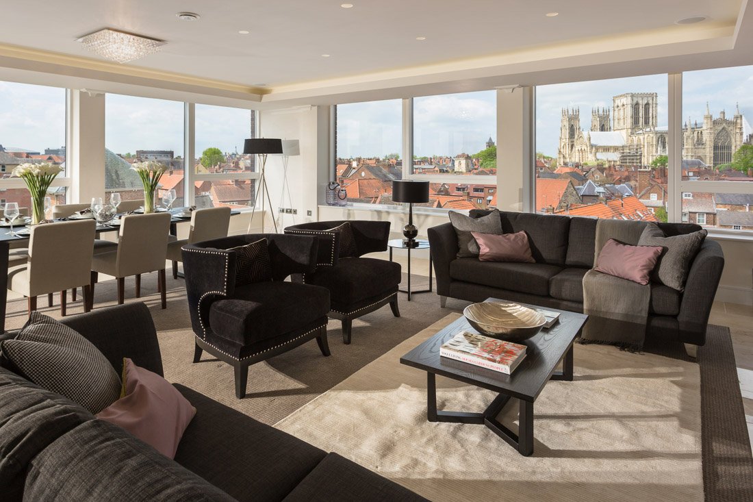  well lit modern apartment living room with panoramic windows looking at York city centre and the minster, furnished with dark purple/brown sofas and arm chairs 