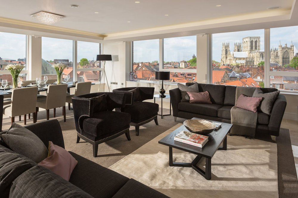  well lit modern apartment living room with panoramic windows looking at York city centre and the minster, furnished with dark purple/brown sofas and arm chairs 