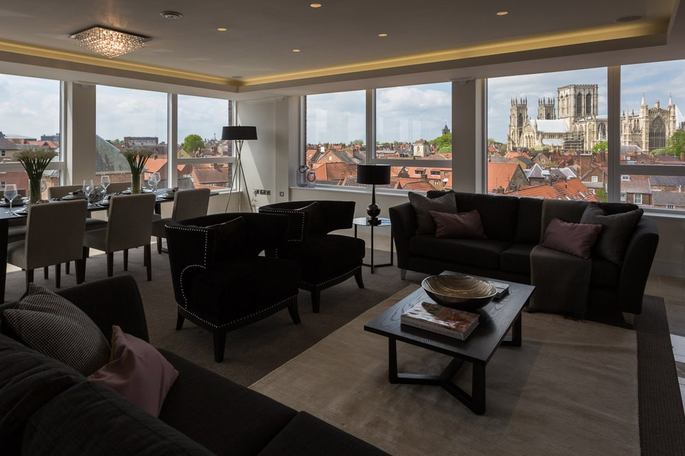  dark badly lit modern apartment living room with panoramic windows looking at York city centre and the minster, furnished with dark purple/brown sofas and arm chairs 