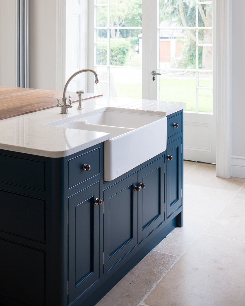  detail of sink on island, beige tile flooring,  navy units, white countertop, porcelain sink and French doors leading out to garden 