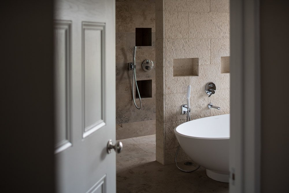  detail image through bathroom door focussed on bath and shower taps, freestanding curved bath, textured beige tiles  