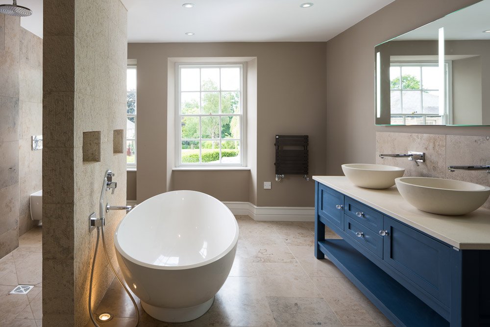  one point perspective of bathroom with large dark blue double sink unit, freestanding curved bath and glimpse into shower, pale beige tiles on floor and walls 