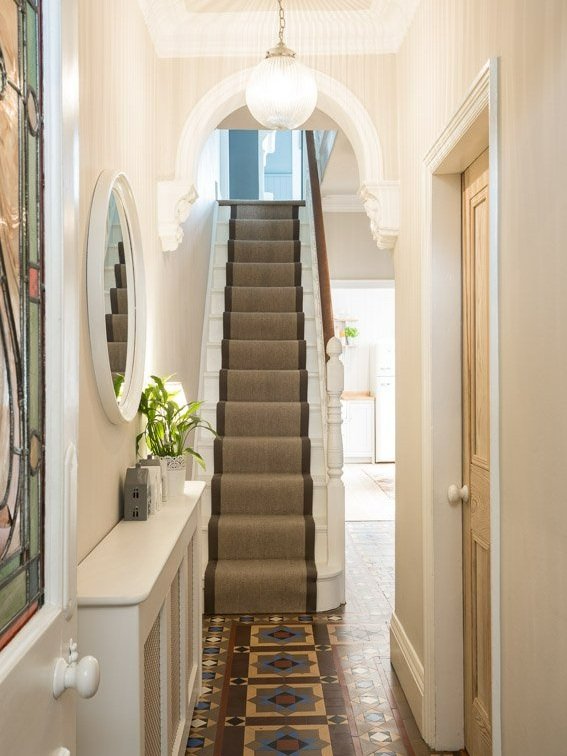  well lit hallway with stained glass door, traditional tile flooring, white wooden stairs with carpet runner, kitchen can just be seen in the background  