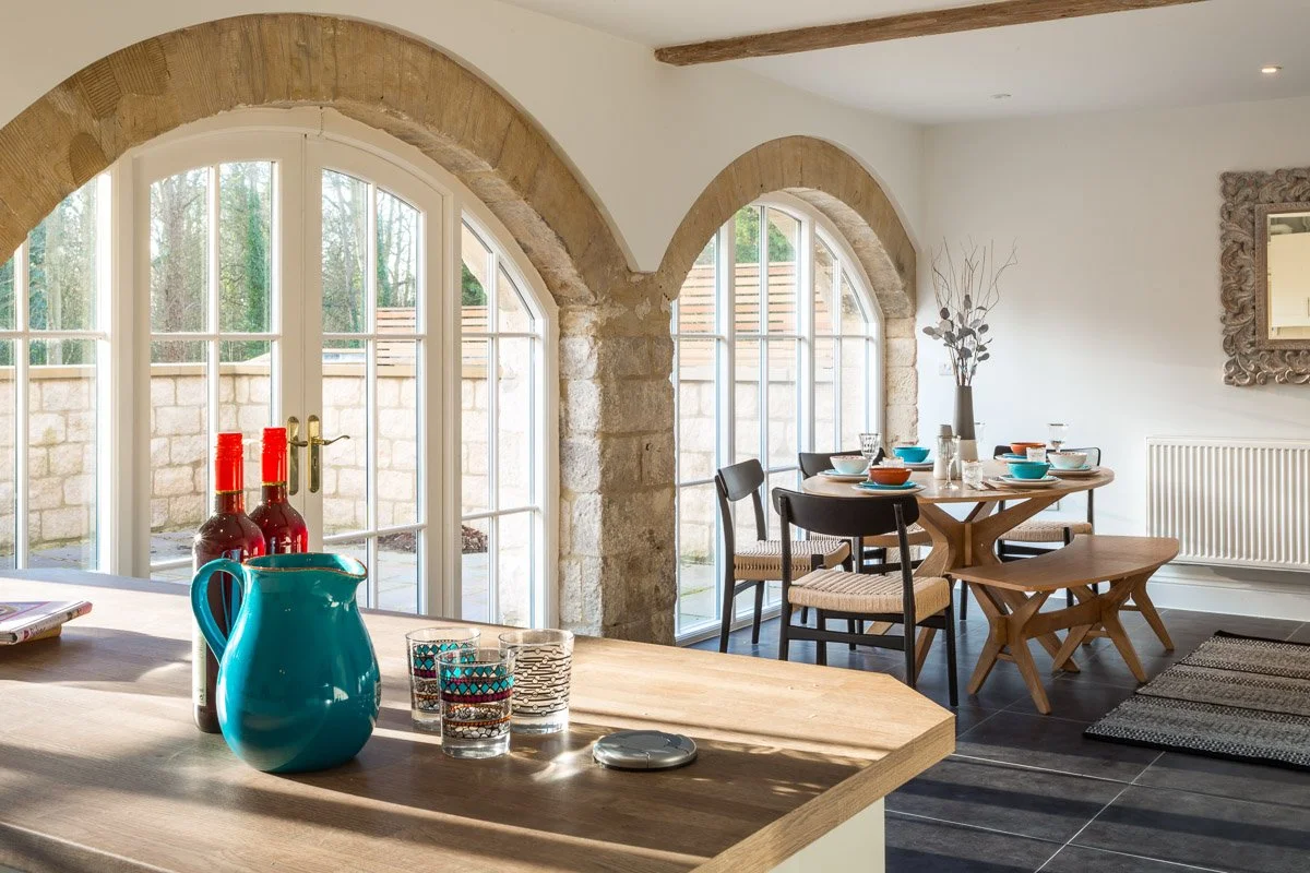 large kitchen diner with arched windows, looking over wooden kitchen island with jug and glasses of water, large dining table with bench seat in background, dark tile flooring, white walls