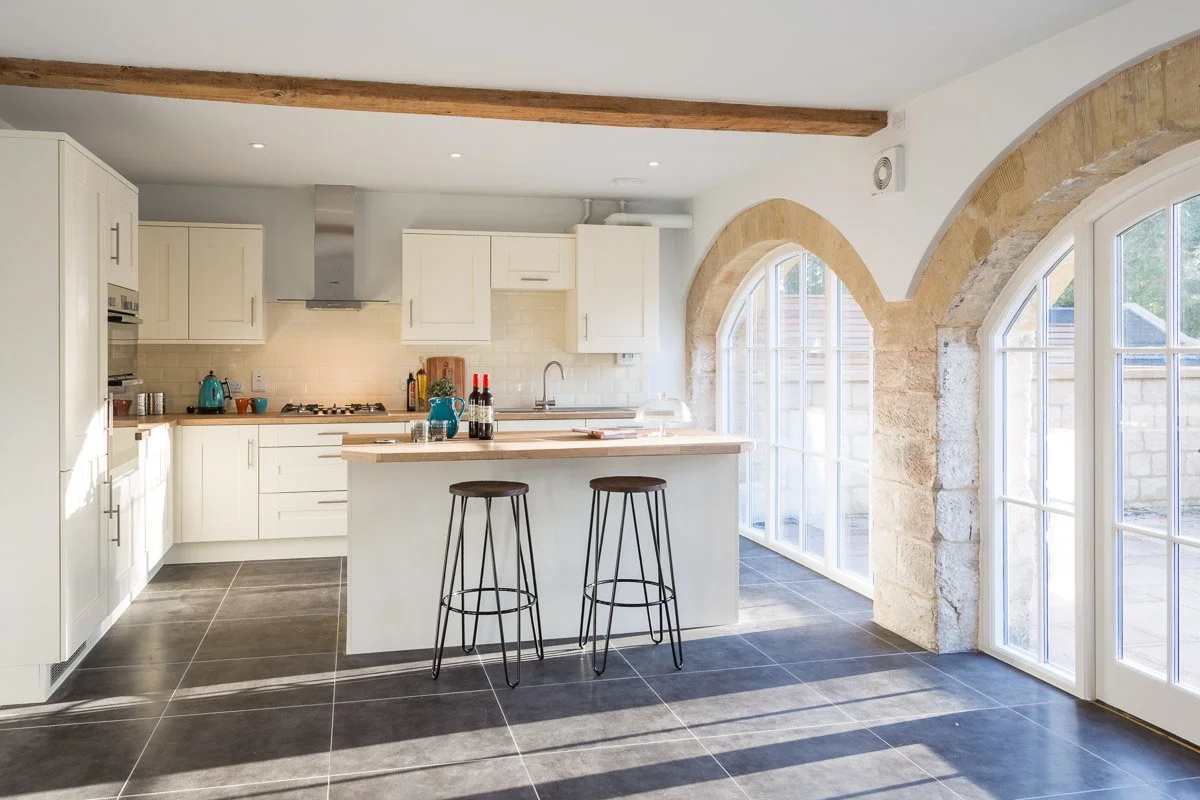 large kitchen with white units, wooden worktops, large arched windows, two metal barstools