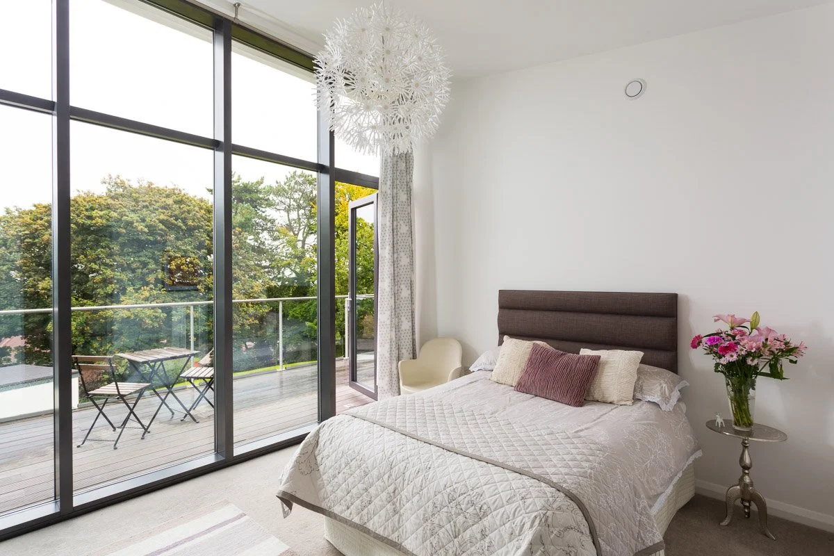 high ceiling bedroom with white walls, full height glass window, beige bed, balcony beyond glass wall