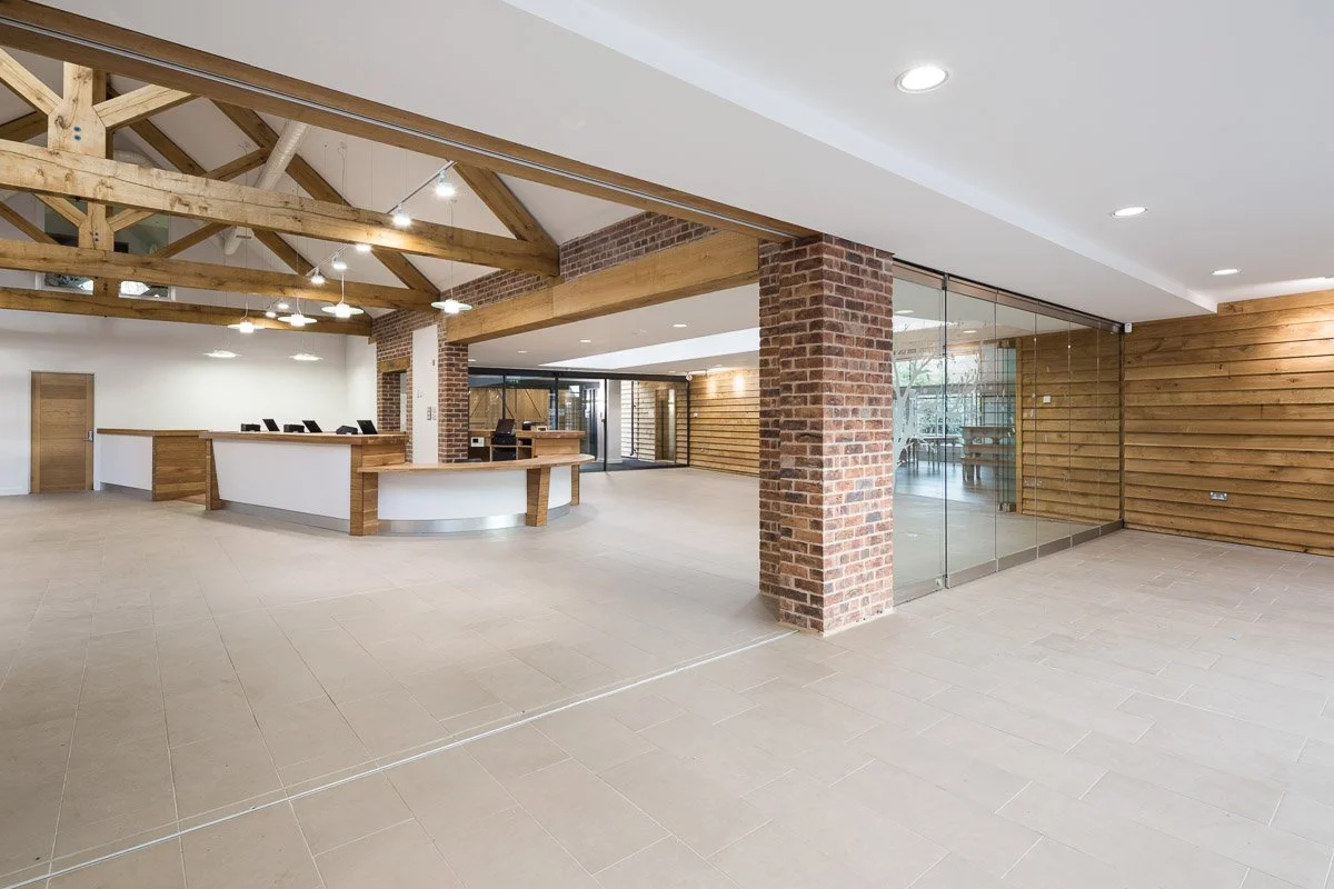 reception room, high beamed ceiling with pale tiled flooring, large curved white and wooden desk, brick pillar in centre of room