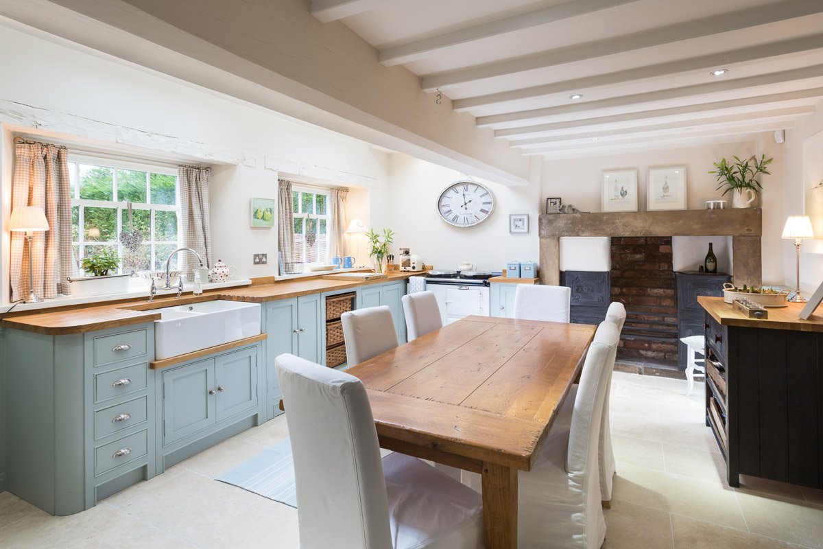 farmhouse kitchen with low white beamed ceiling, pale blue units, wooden counter tops, wooden dining table, beige tile floor