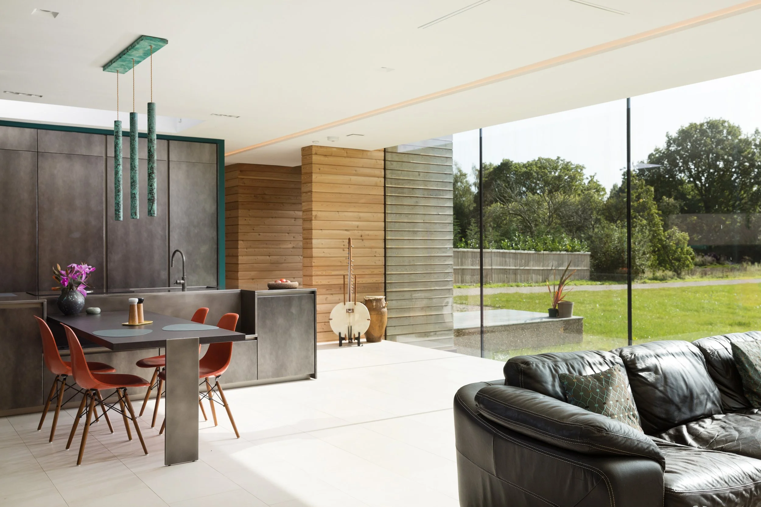 large modern kitchen/dining/family room with glass wall and wood panelled wall, dark dining table and leather sofas looking out to the garden beyond