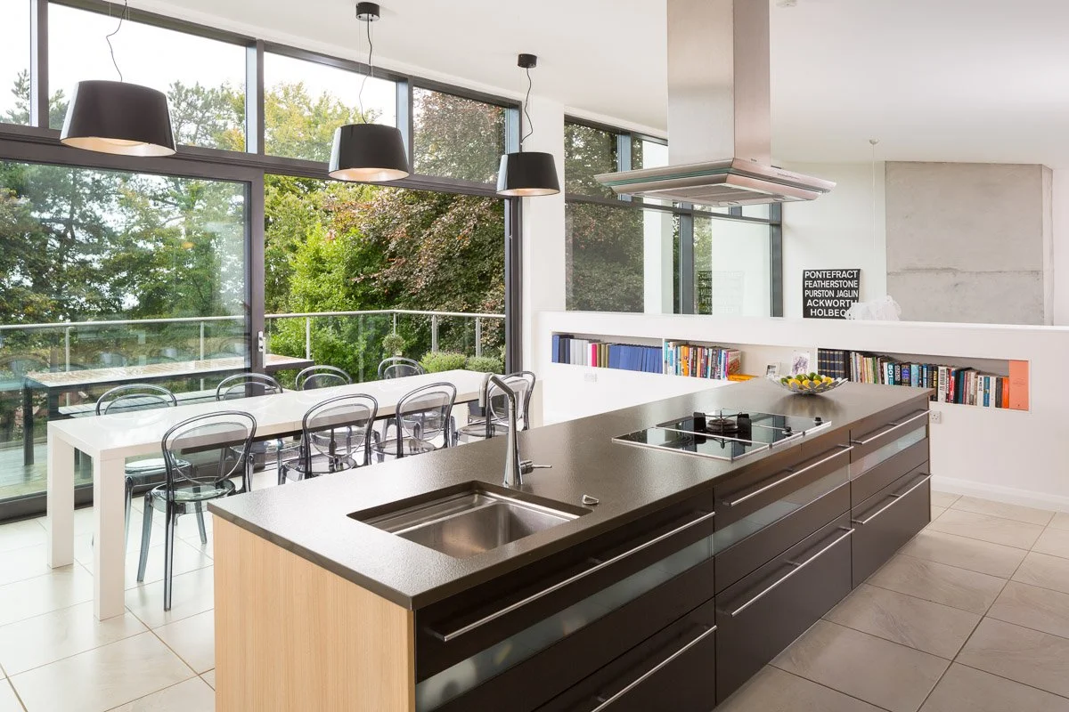 modern kitchen with tile flooring, dark units, pendant lights, white dining table, full height glass wall with balcony beyond 