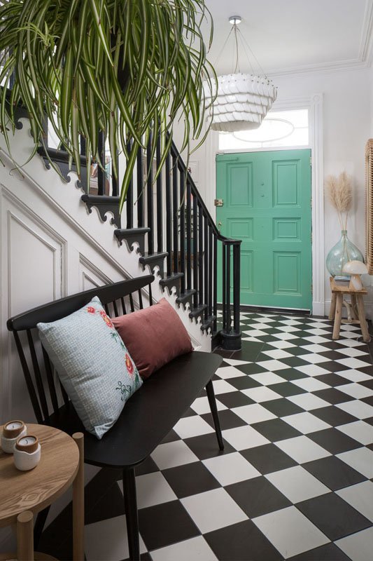 portrait image of hallway with mint green front door, black and white check tile flooring, black bench seat and spider plant hanging from stairs on the right