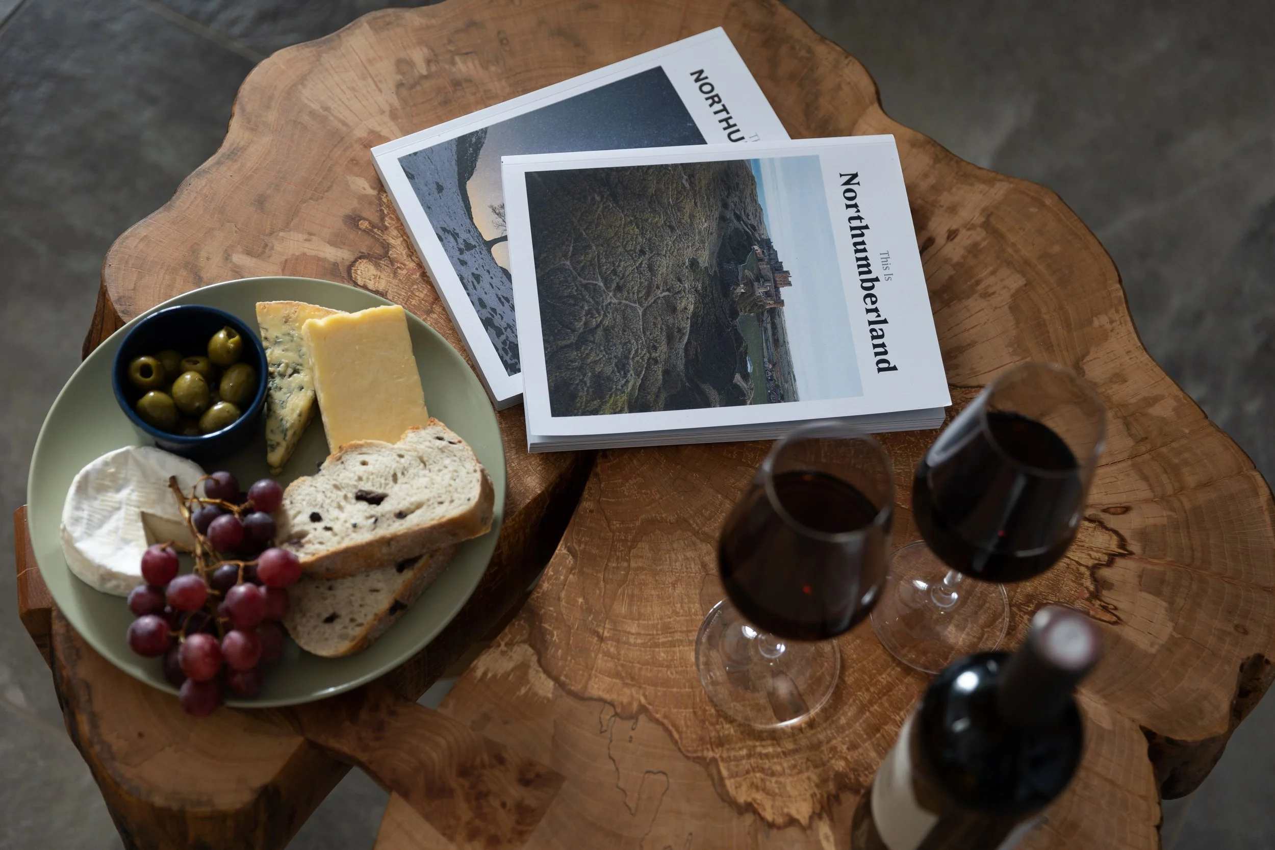unique wooden table with two glasses of red wine, plate of mixed cheeses and grapes and two books about Northumberland 