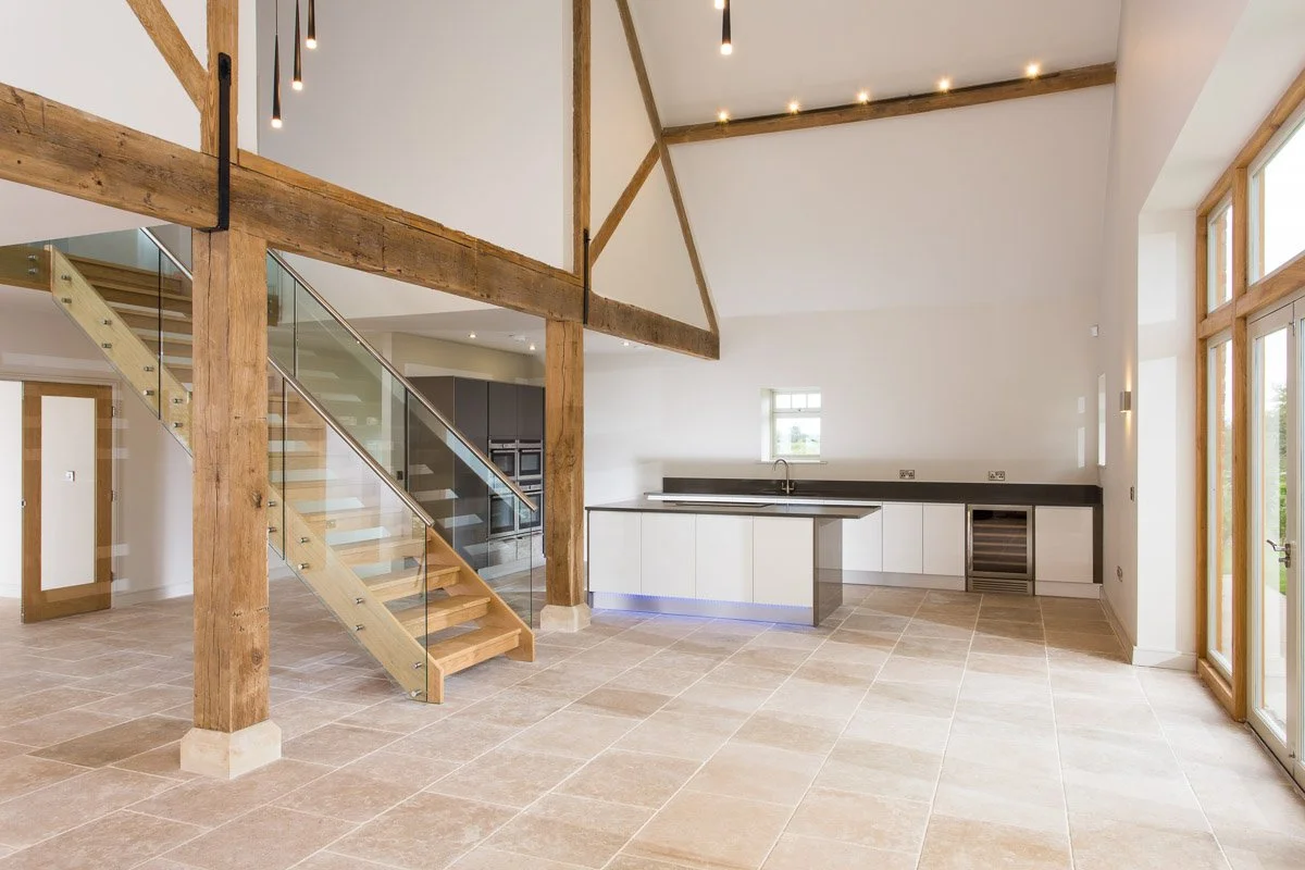 open plan double height ceiling kitchen diner, wooden stair case with glass banister, wooden pillars, white kitchen units with dark counter tops
