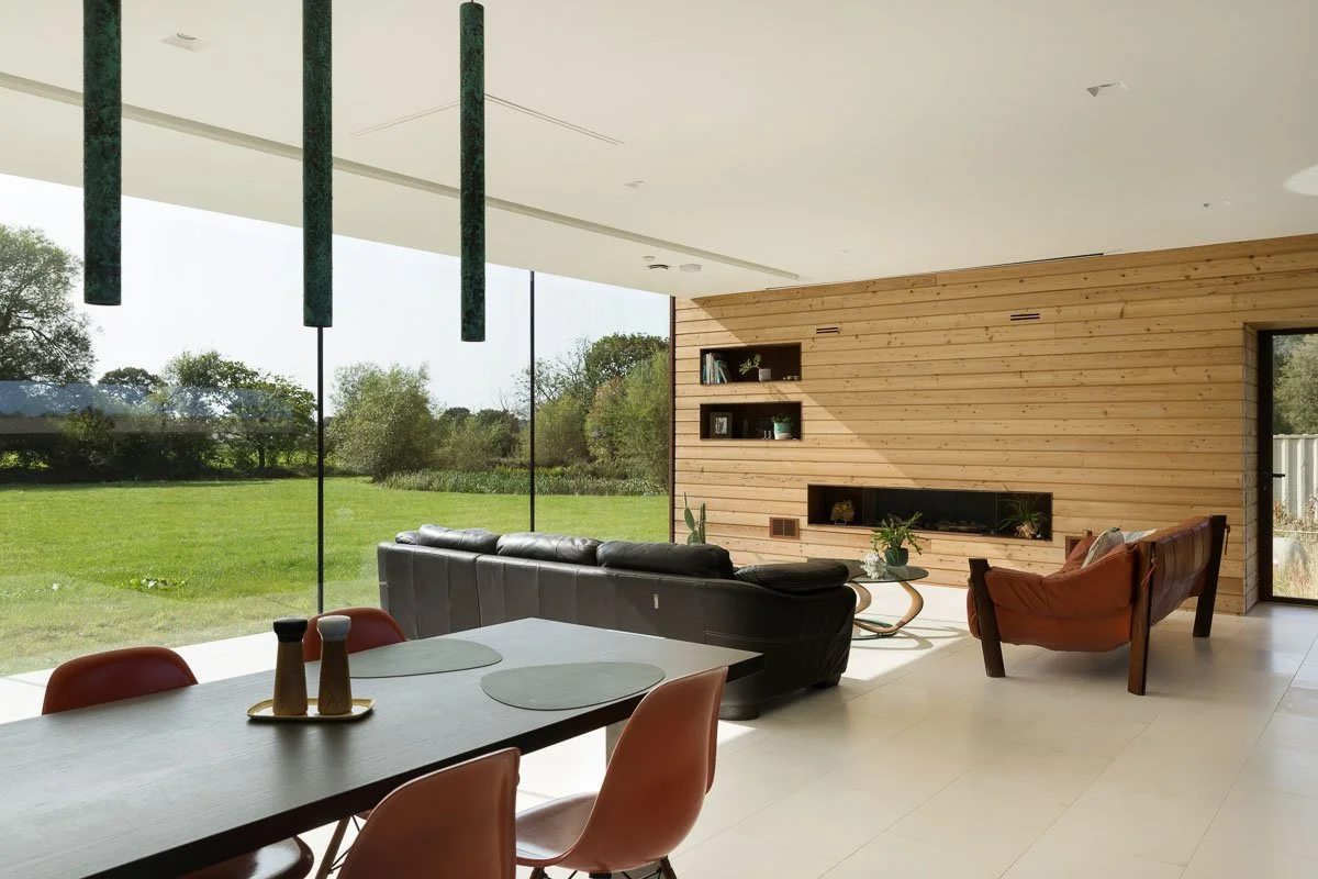 large modern dining/family room with glass wall and wood panelled wall, dark dining table and leather sofas looking out to the garden beyond  