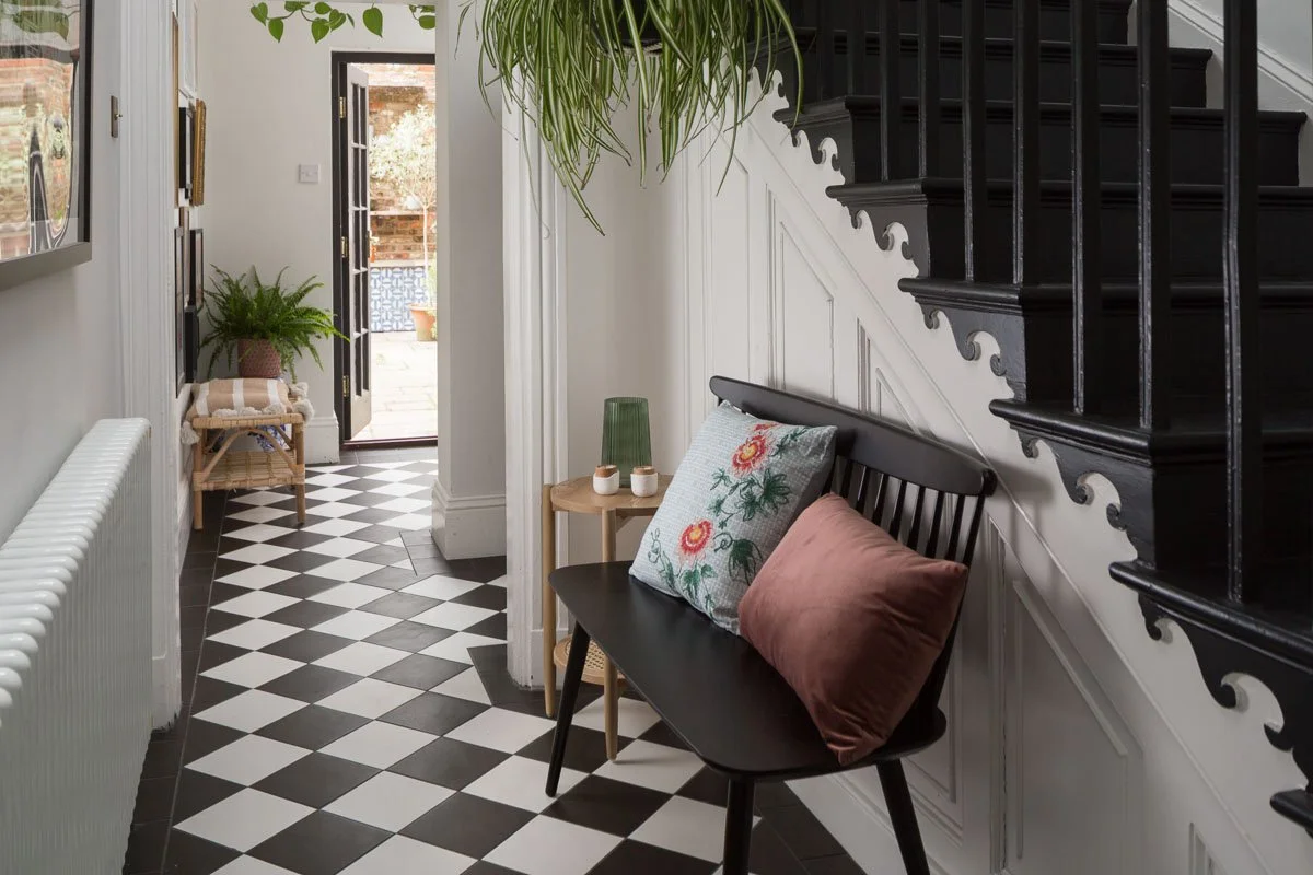 hallway with black and white check flooring, black stair case, black bench seat, spider plant hanging from stairs on left, glimpse into room beyond