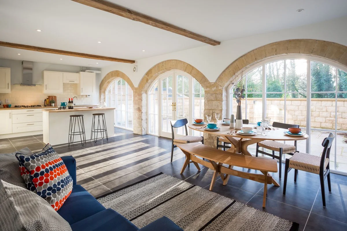 south facing kitchen/diner with high ceilings and large arched windows, round wooden kitchen table, blue sofa and two metal stools at the kitchen island, on a sunny day with light streaming in through the large arched windows