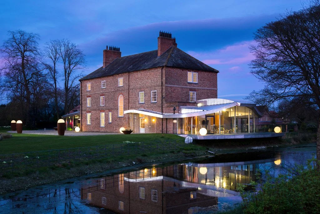 Large manor house shot at dusk with glowing yellow lights from the windows, reflected in the stream running alongside the house