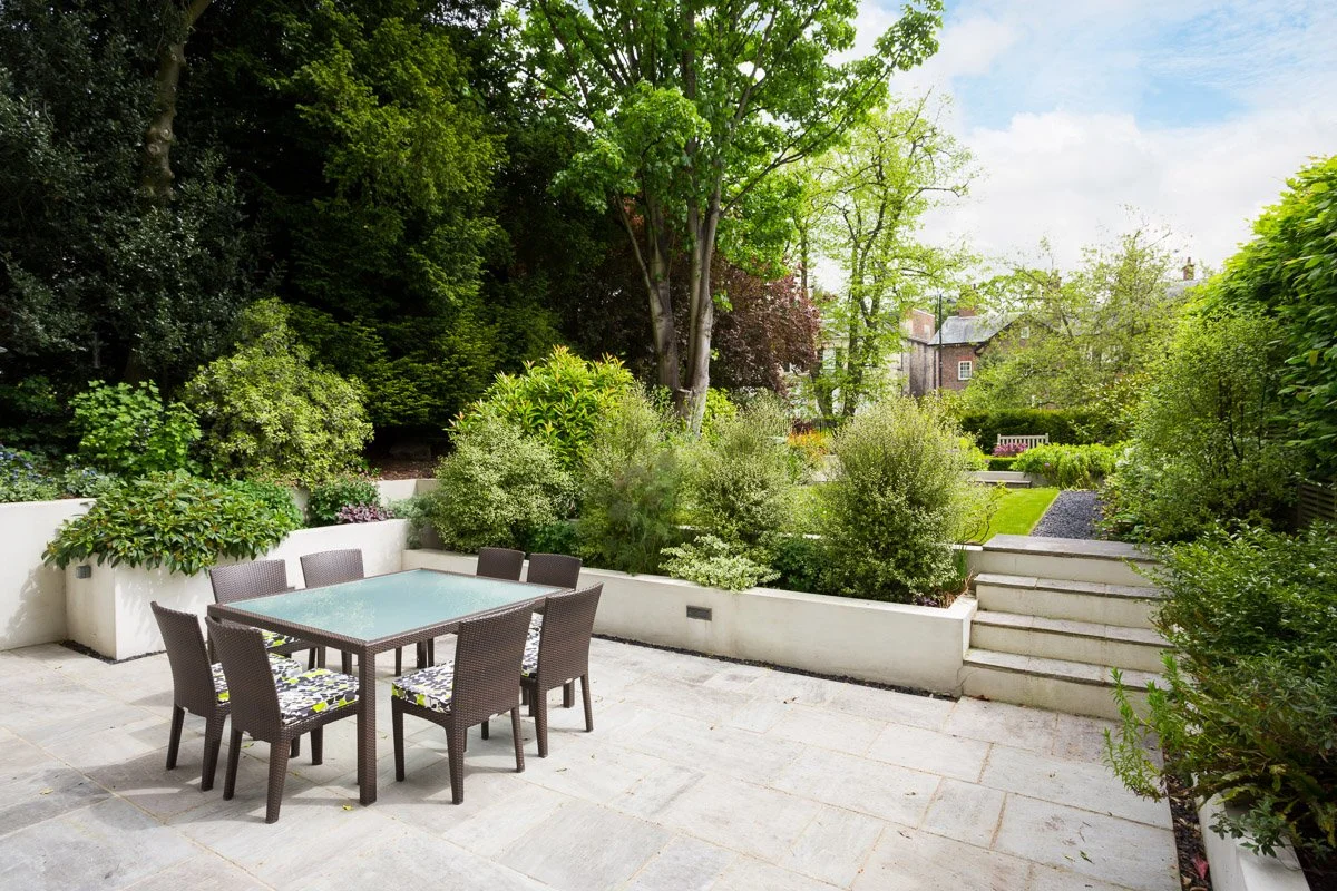 patio area with glass table and woven chairs, beyond neat raised beds and a grass law up a couple of stone steps, surrounded by tall trees and hedges