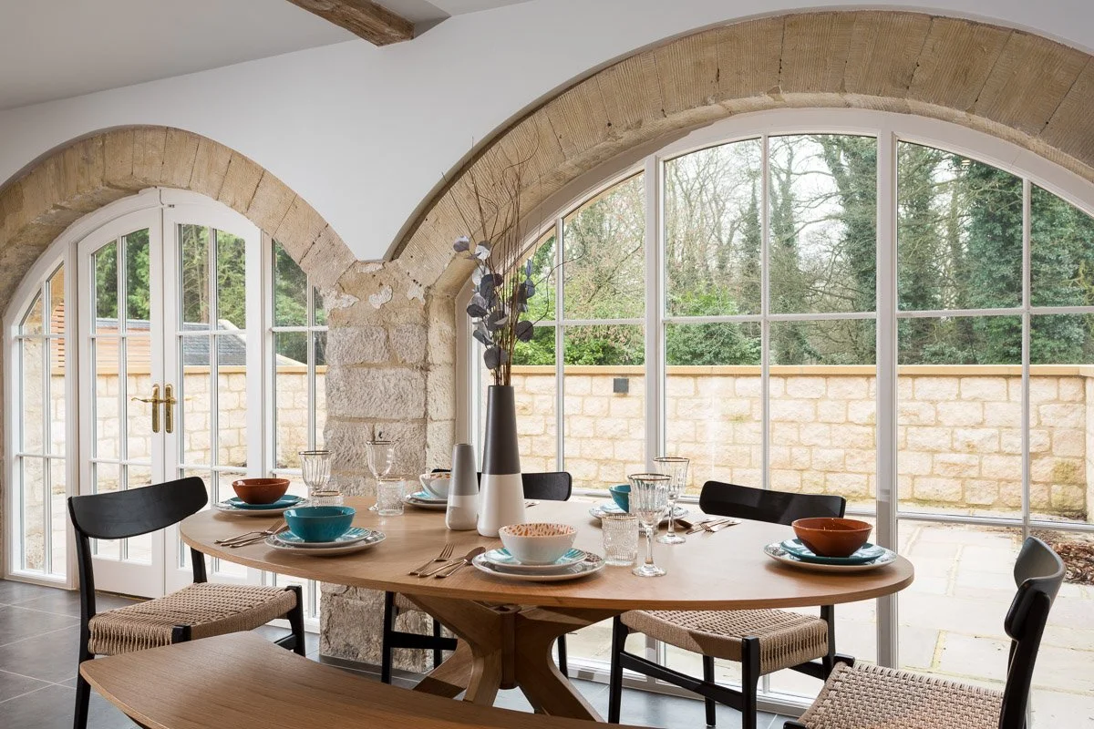 detail of dining table, wooden table with plates and cutlery, large arched windows behind 