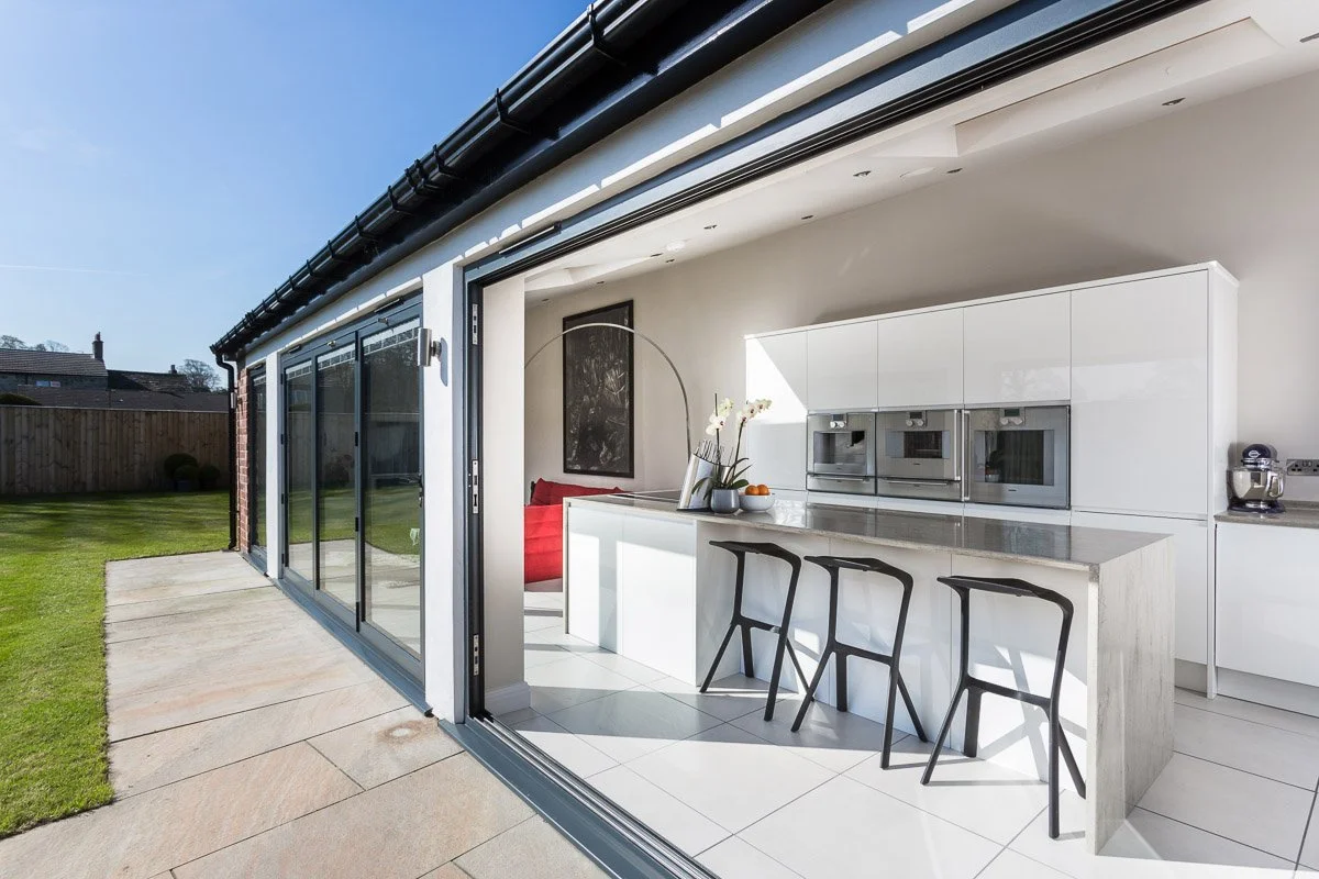 open plan kitchen diner photographed from outside with bifolds open, white kitchen units with black metal stools 