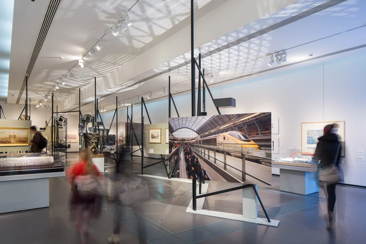 inside exhibit 'destination stations' several pictures of york rail history under show lighting, blurred people walk by