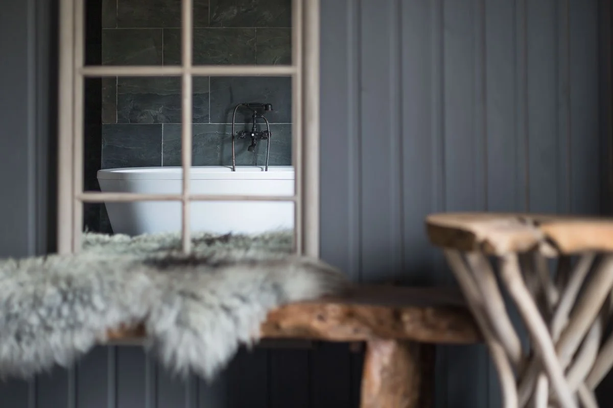 detail shot of blue outside wooden walls, wooden bench seat with sheepskin rug, glimpse through window at large curved free standing bath