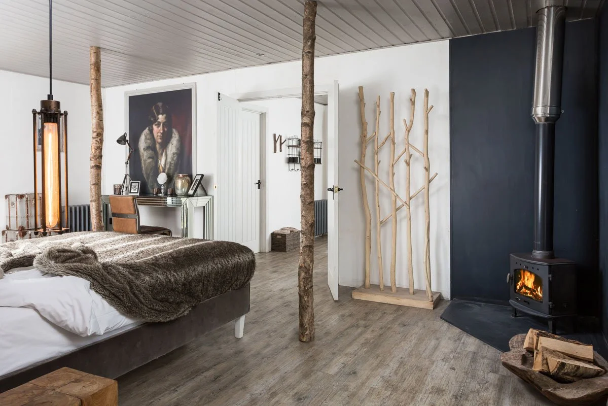 large holiday home bedroom with wooden flooring, navy blue wall, log burner in corner, fur blanket on bed