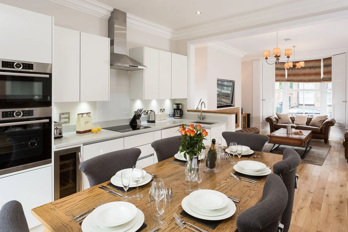 looking across wooden dining table towards kitchen and living room, white units, wooden flooring, large leather sofas 