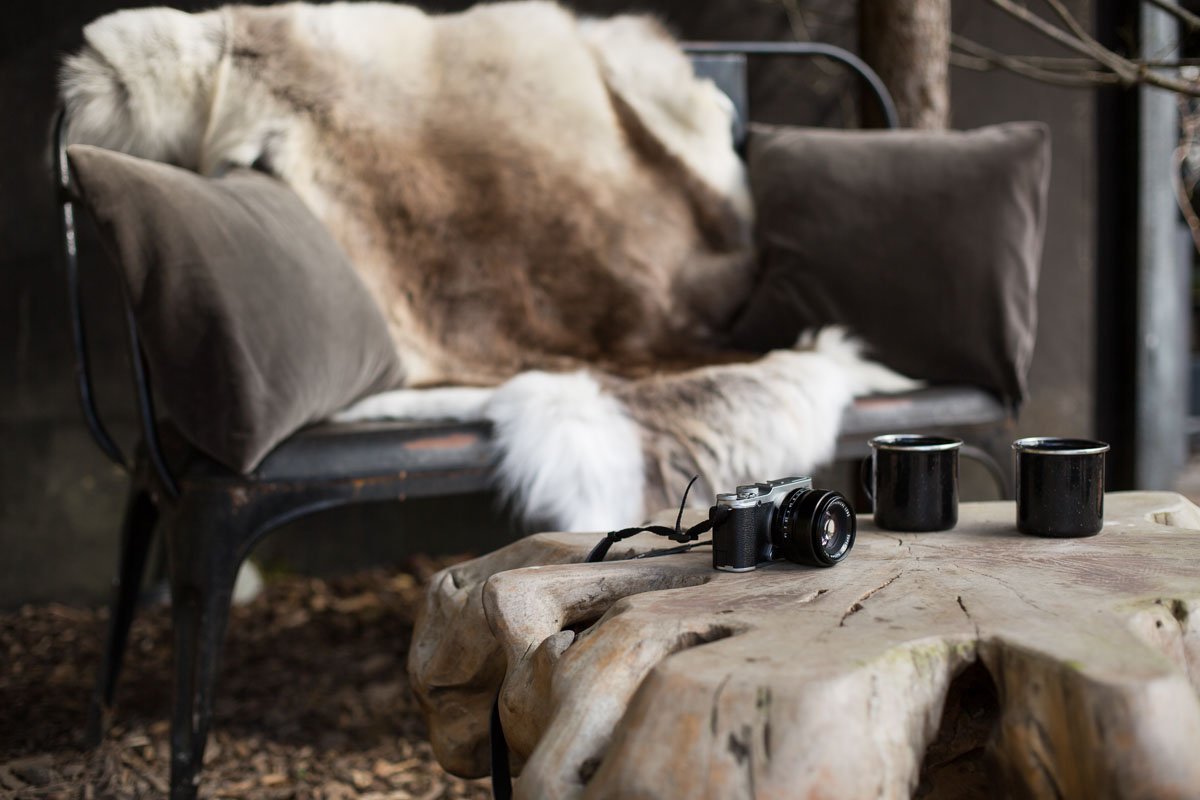 large dark grey armchair with sheepskin behind wooden tree trunk table with film camera and two mugs on