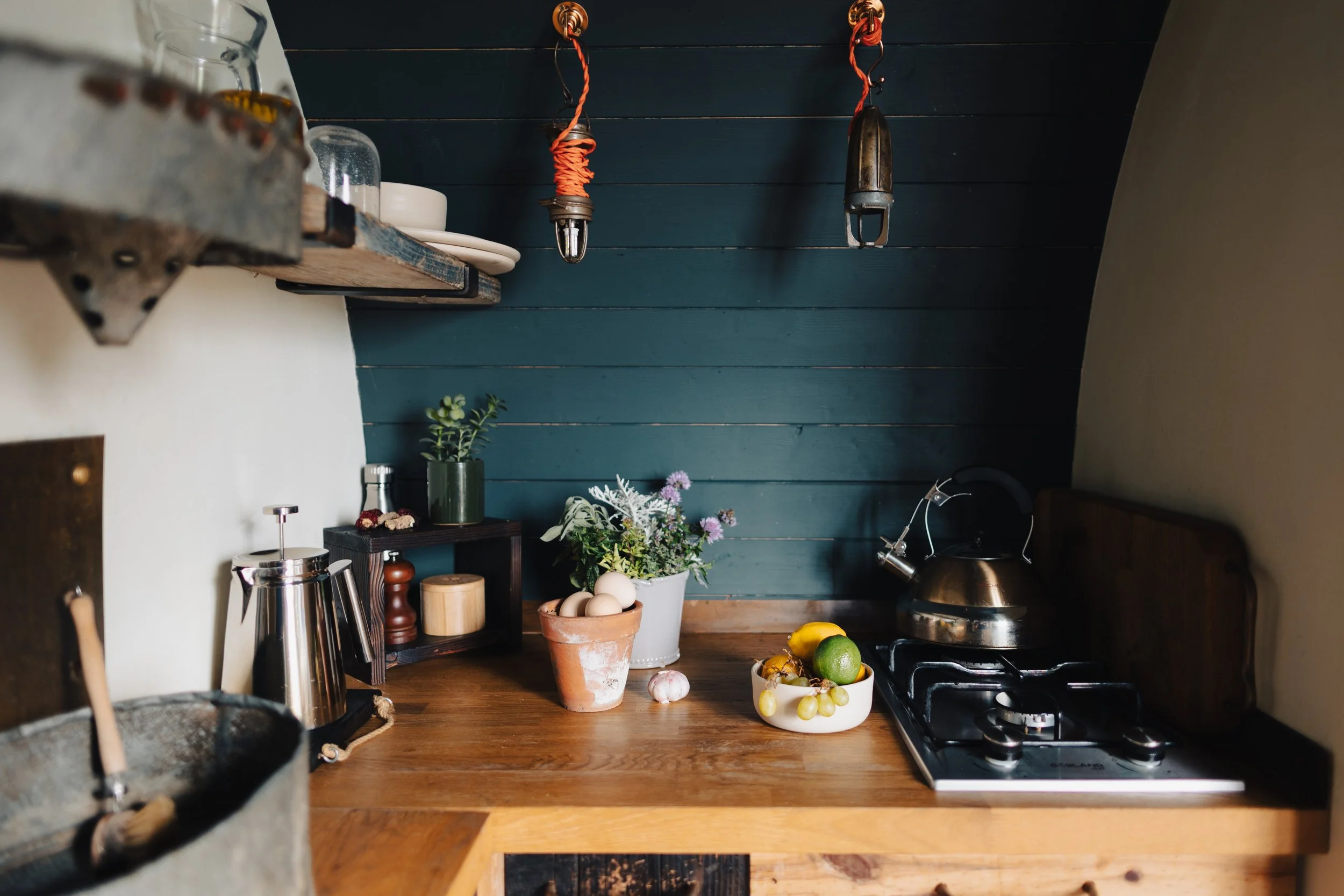 kitchen nook in shepherds hut with wooden worktops, dark teal wall, pendant lights and cooking utensils