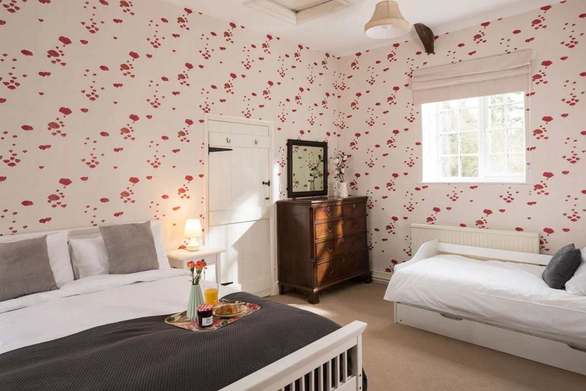 detail of corner of bedroom,  looking across bed with breakfast tray on, small white sofa bed in right hand corner, wooden draw unit against red and white wallpapered wall 