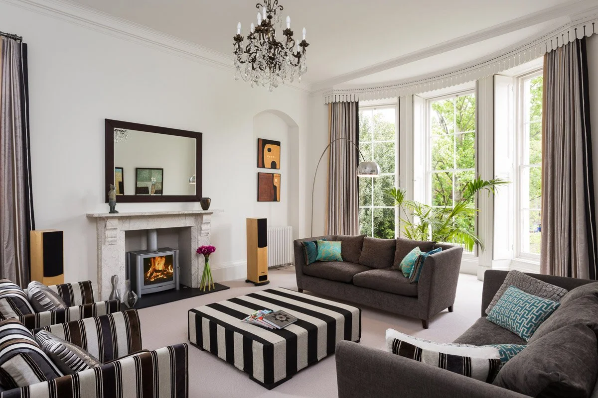 large high ceiling living room with white walls, grey sofas, black and white striped arm chairs and ottoman, fire place with log burner, large bay window