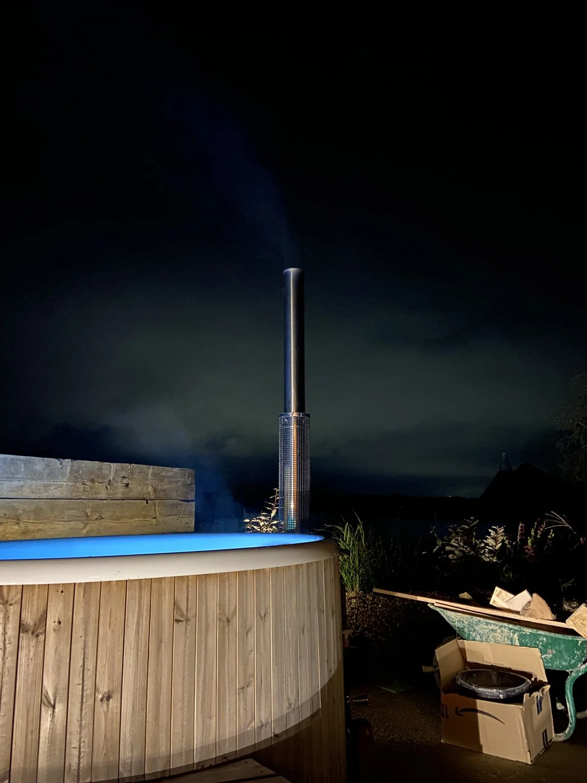 An outdoor hot tub with wooden paneling, illuminated with blue lighting, situated near gardening tools and boxes at night against a dark cloudy sky.