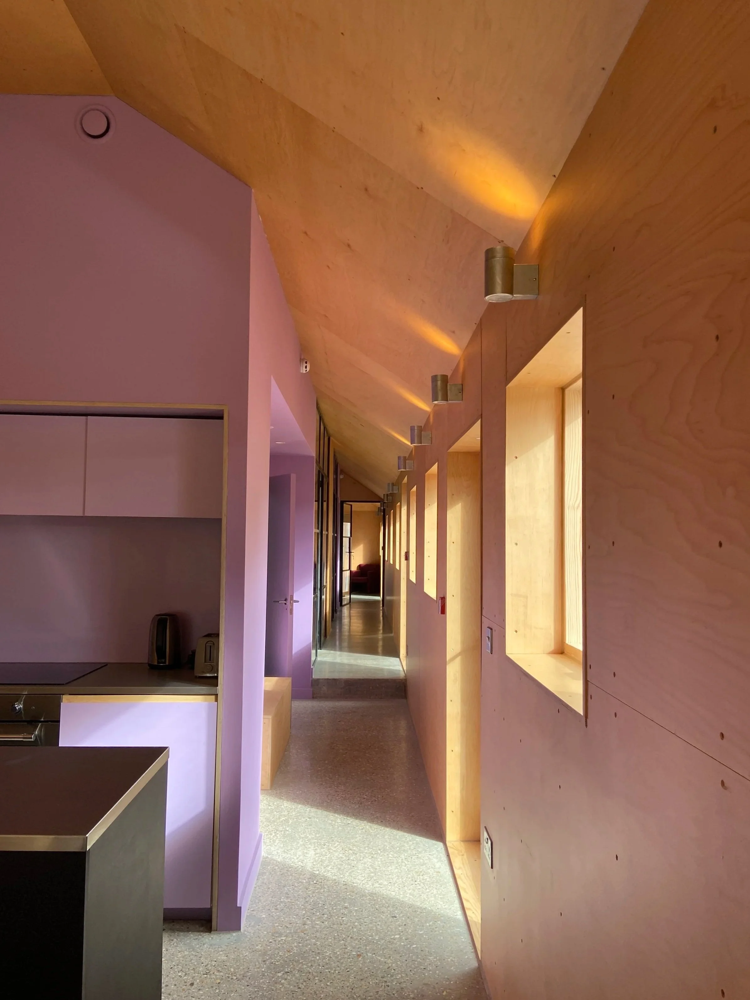 Interior corridor with wooden wall, ceiling, and orange wall lights, leading to a room in the distance.