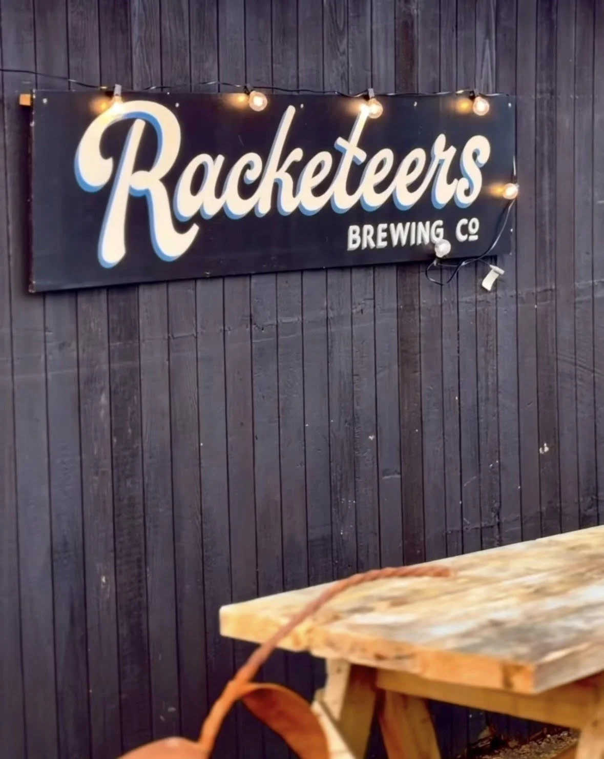 Sign for Racketters Brewing Co with string lights hanging above, mounted on a dark wooden wall.