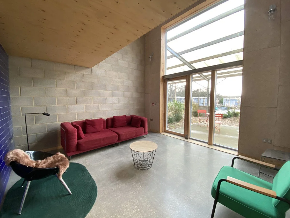 Modern living room with a red sofa, a green armchair, a small round coffee table, and a black lounge chair with a fur throw. Large windows let in natural light and reveal an outdoor patio with red chairs.