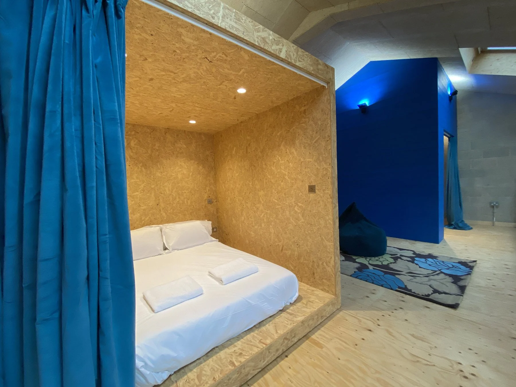 A room with a bed under a wooden canopy, blue curtains on the left, and a separate blue wall section with a blue bean bag and a patterned rug on the right.