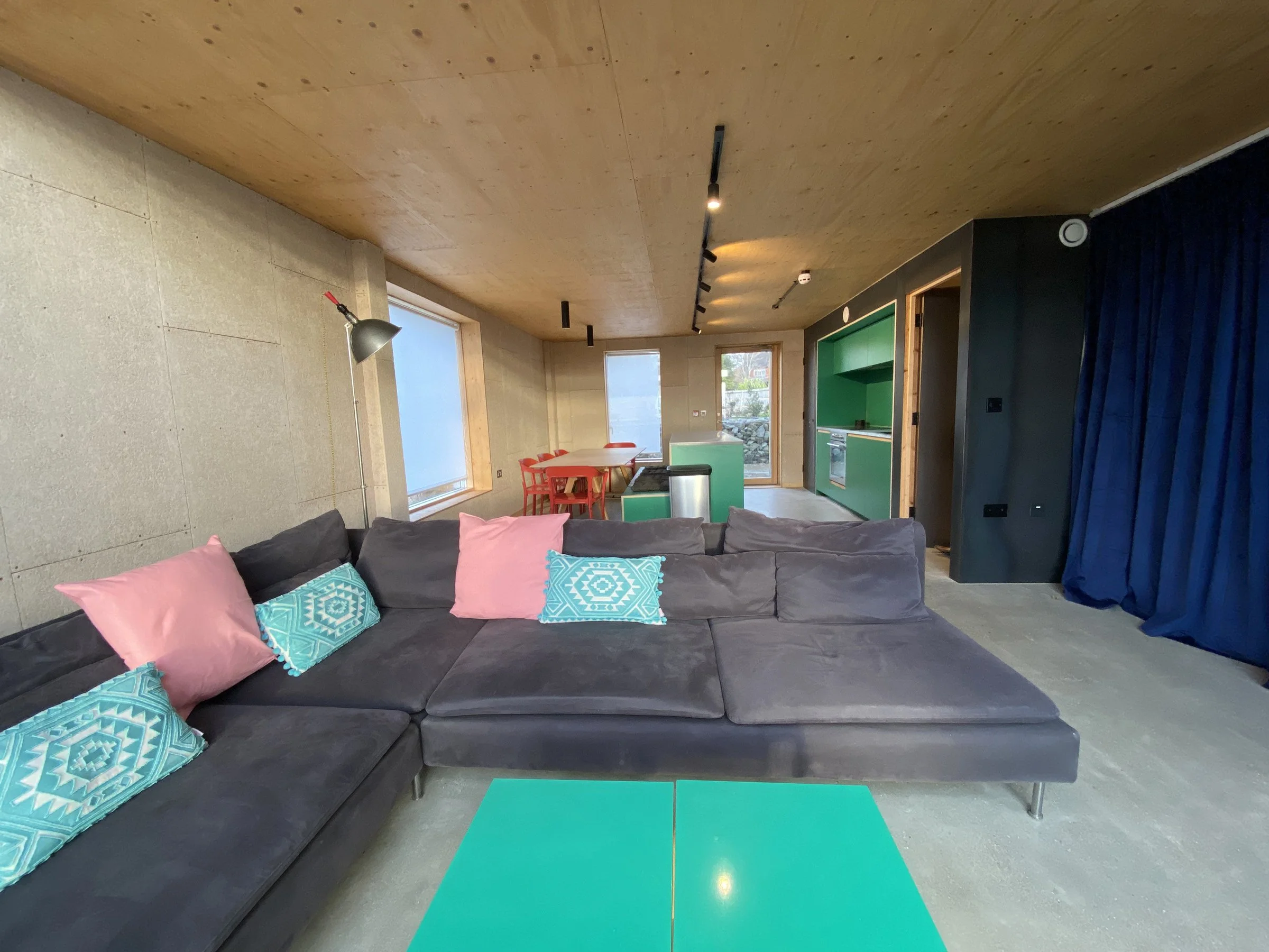 Living room with dark gray sectional sofa, pink and blue decorative pillows, blue coffee table, wooden ceiling, large windows, green kitchen cabinetry, dining table with red chairs, and blue curtains.
