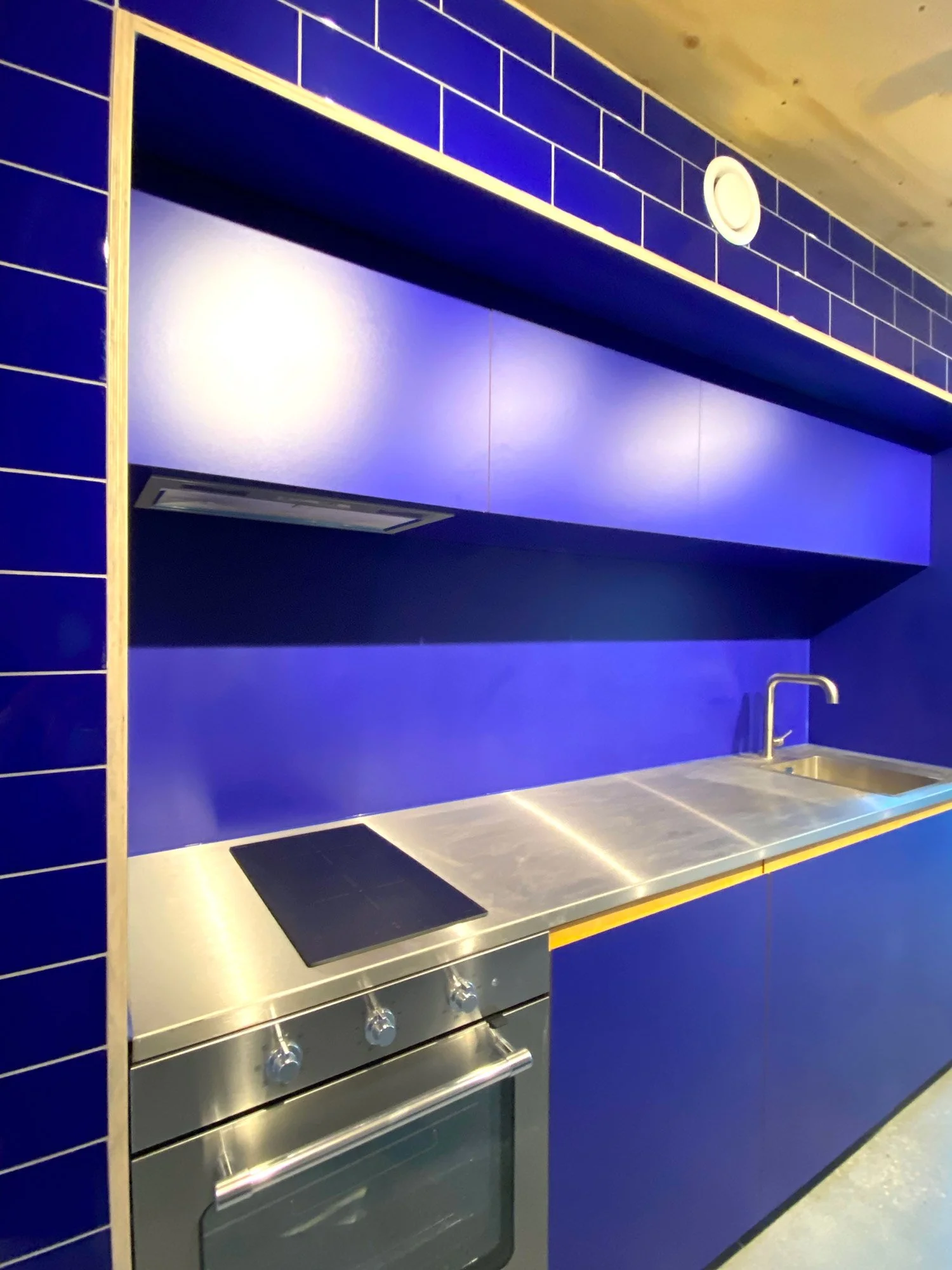 A modern kitchen with blue cabinets, a stainless steel countertop, a built-in stove, and a blue wall with a ventilation hood.