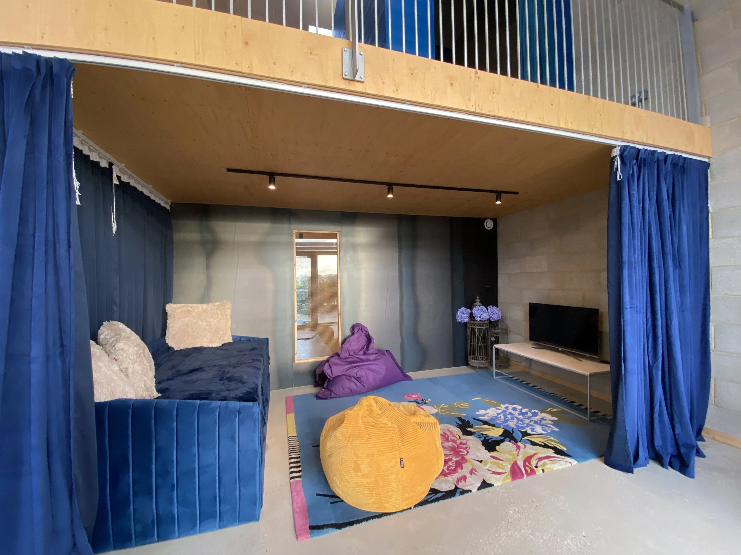 Living room with blue curtains, multicolored patterned rug, purple bean bag, yellow pouf, blue velvet sofa with plush pillows, TV on a sleek stand, vase with purple hydrangeas, and a window with a city view.