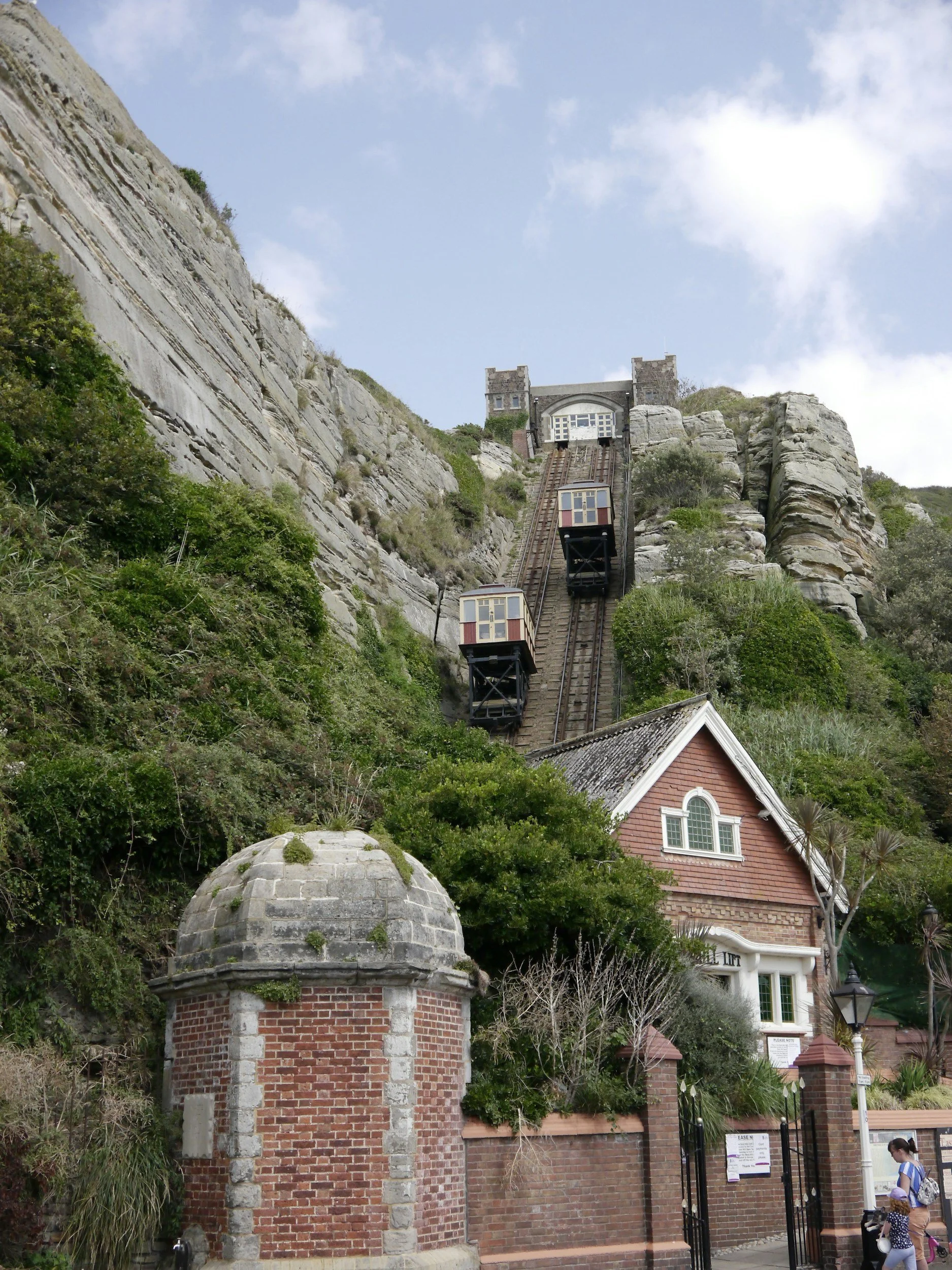 A steep hillside with a funicular railway and two small cabins on the tracks, leading to a building at the top, with greenery surrounding the scene and a person walking near a brick wall in the foreground.