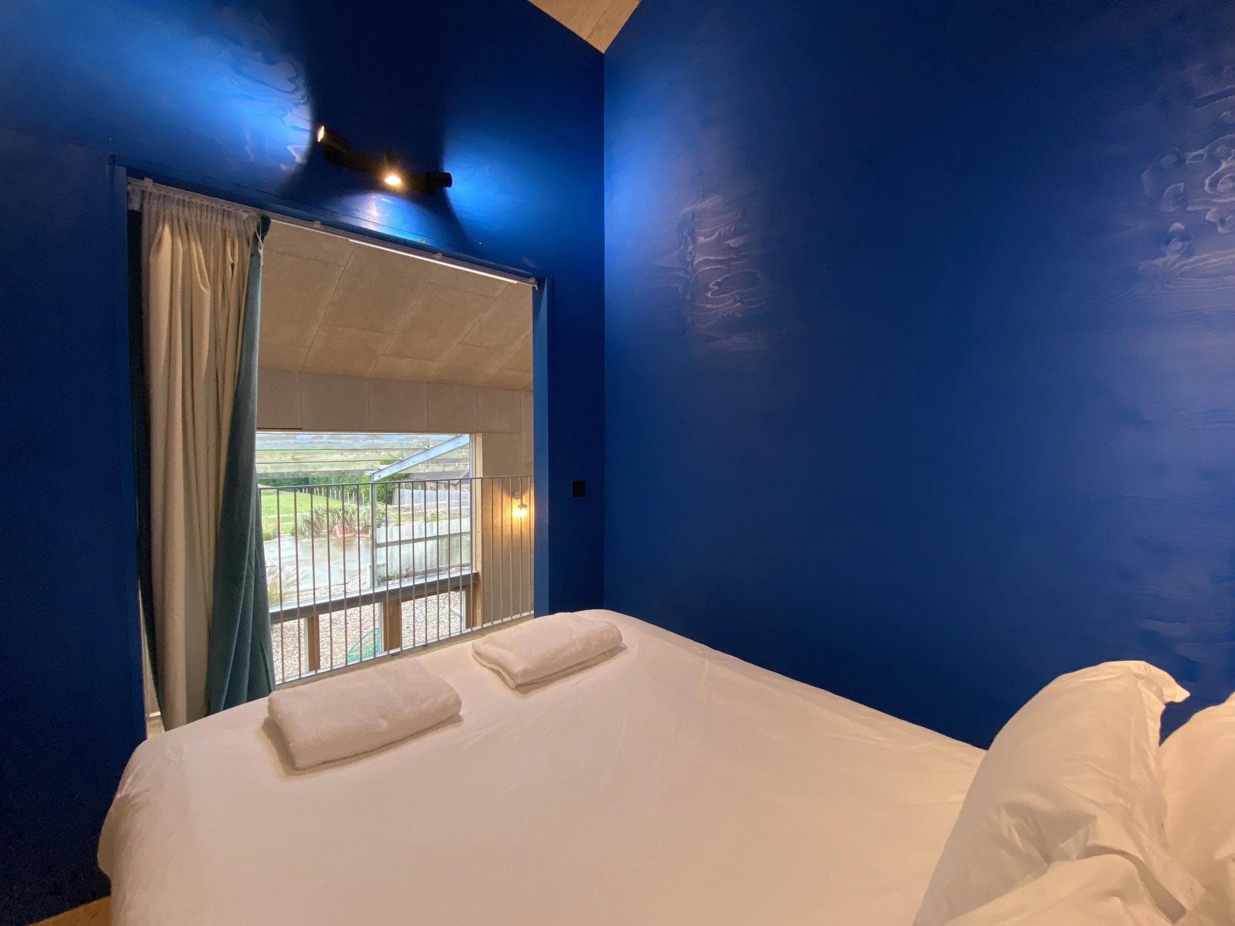 Bedroom with bright blue walls, a large bed with white linens and pillows, and a window with curtains showing an outdoor scene with grass and buildings.