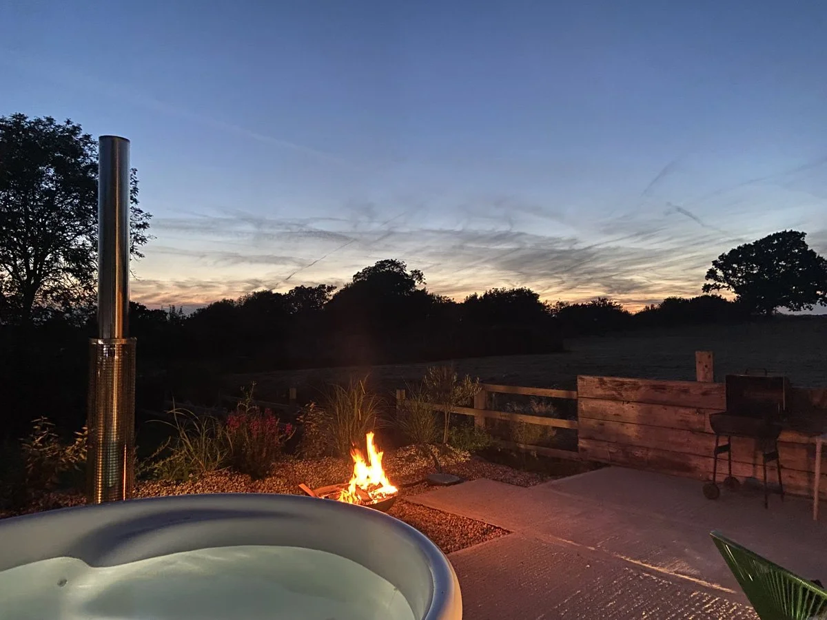 Outdoor scene at dusk with a fire pit, a hot tub in the foreground, a grill on the right, and trees in the background.