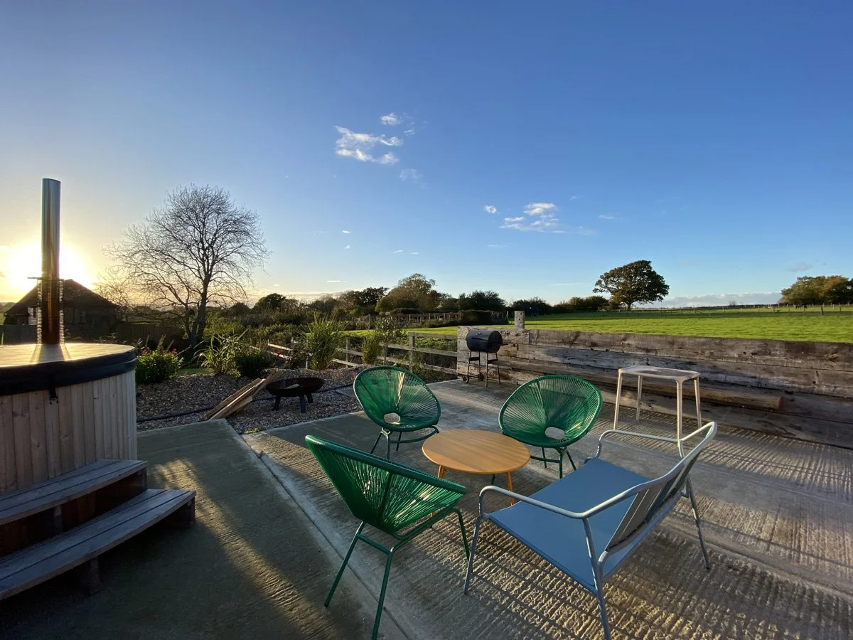 Outdoor patio with green and blue chairs, small wooden table, grill, hot tub, and a view of green fields and trees during sunset.