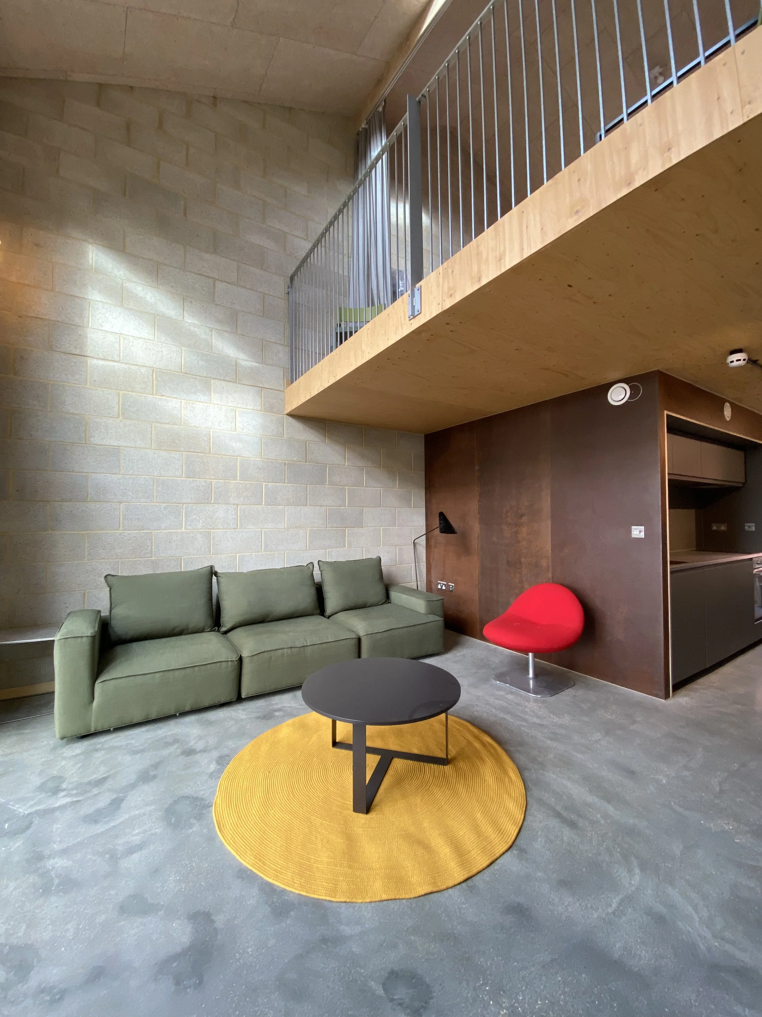 A modern living room featuring a green sofa, a round yellow rug, a black coffee table, a red accent chair, and a textured light-colored brick wall. An upper loft area with a wooden floor and metal railing overlooks the space.