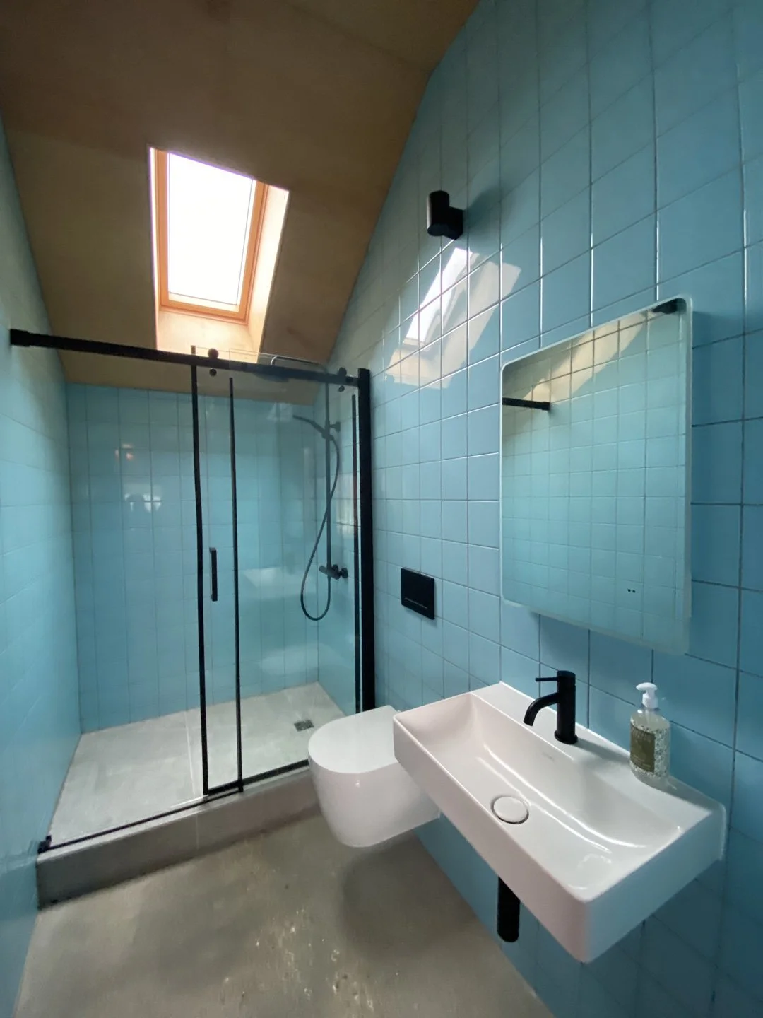 Modern bathroom with blue tiled walls, a glass shower enclosure, a white wall-mounted toilet, a rectangular white sink with a black faucet, a mirror, a black wall lamp, and a skylight.