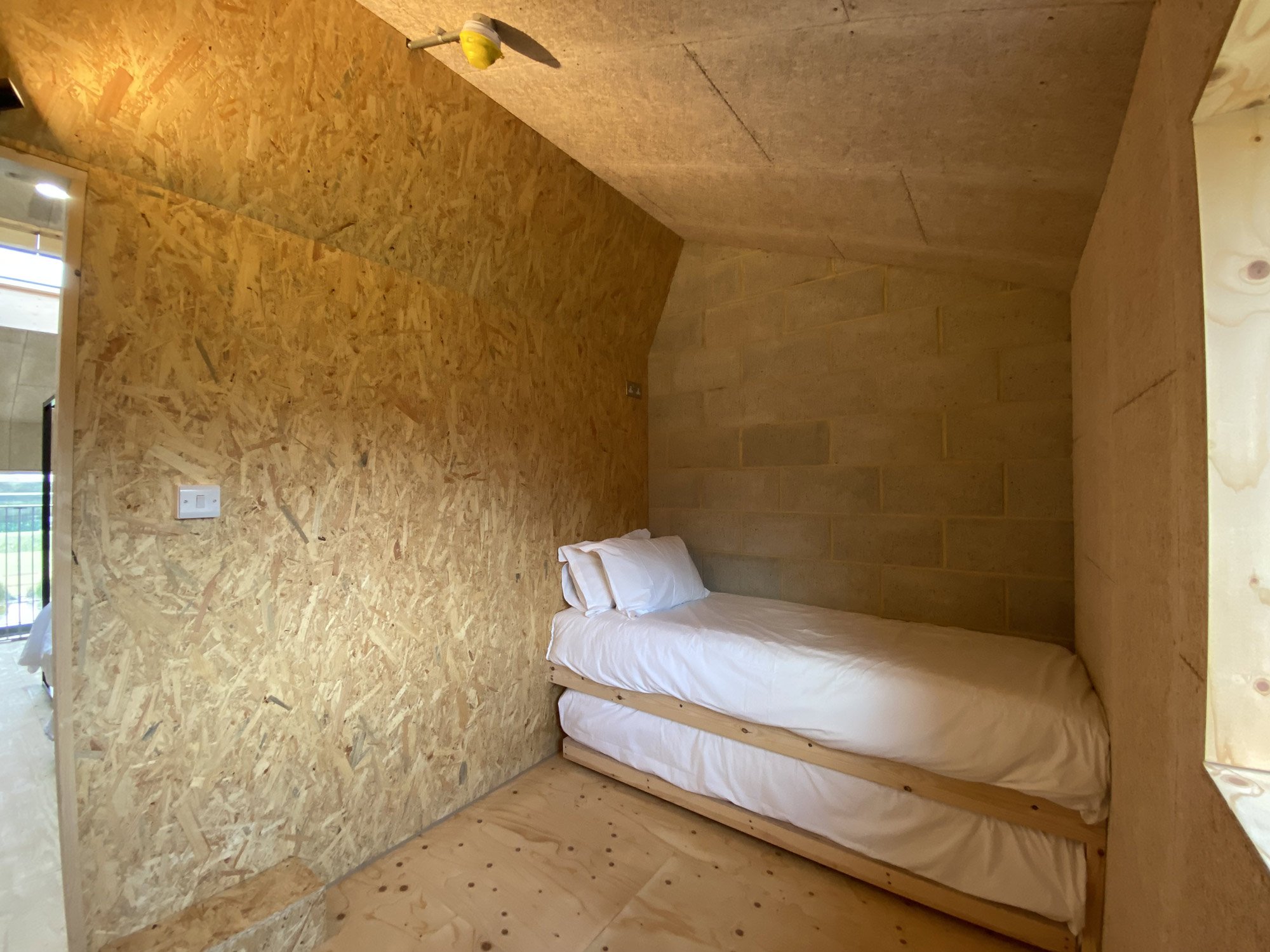 A small sleeping area with a single bed headrest, white bedding, located within a room with plywood and plywood wall panels and a concrete cinderblock wall.